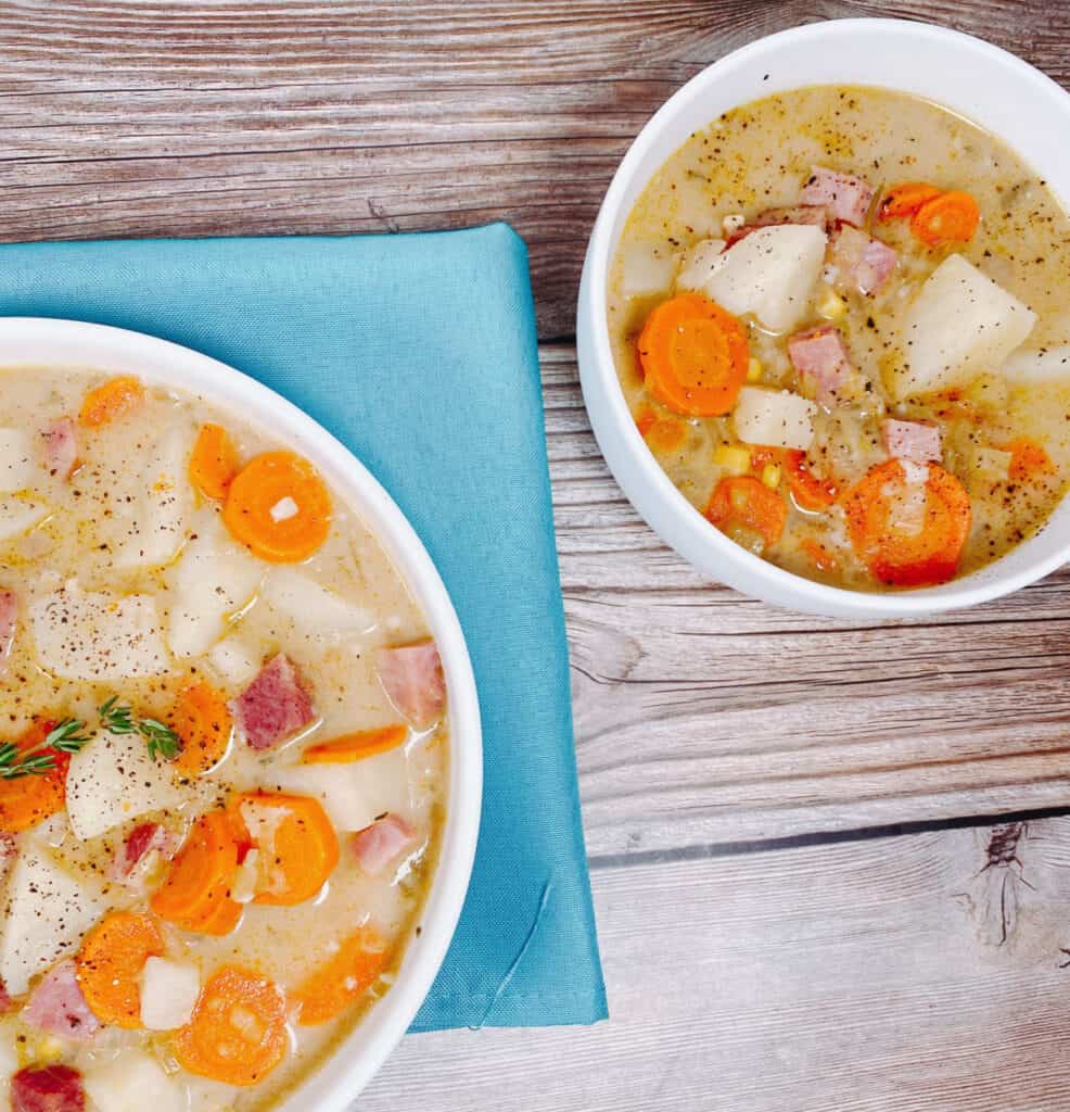 Two bowls of creamy ham and potato soup sit on a wooden background. The bowl on the left sits under a turquoise napkin. In both bowls, pieces of ham, potato, carrots and veggies are prominent over the broth. Both bowls of soup are seasoned with salt and pepper and the left bowl has a sprig of fresh thyme. 