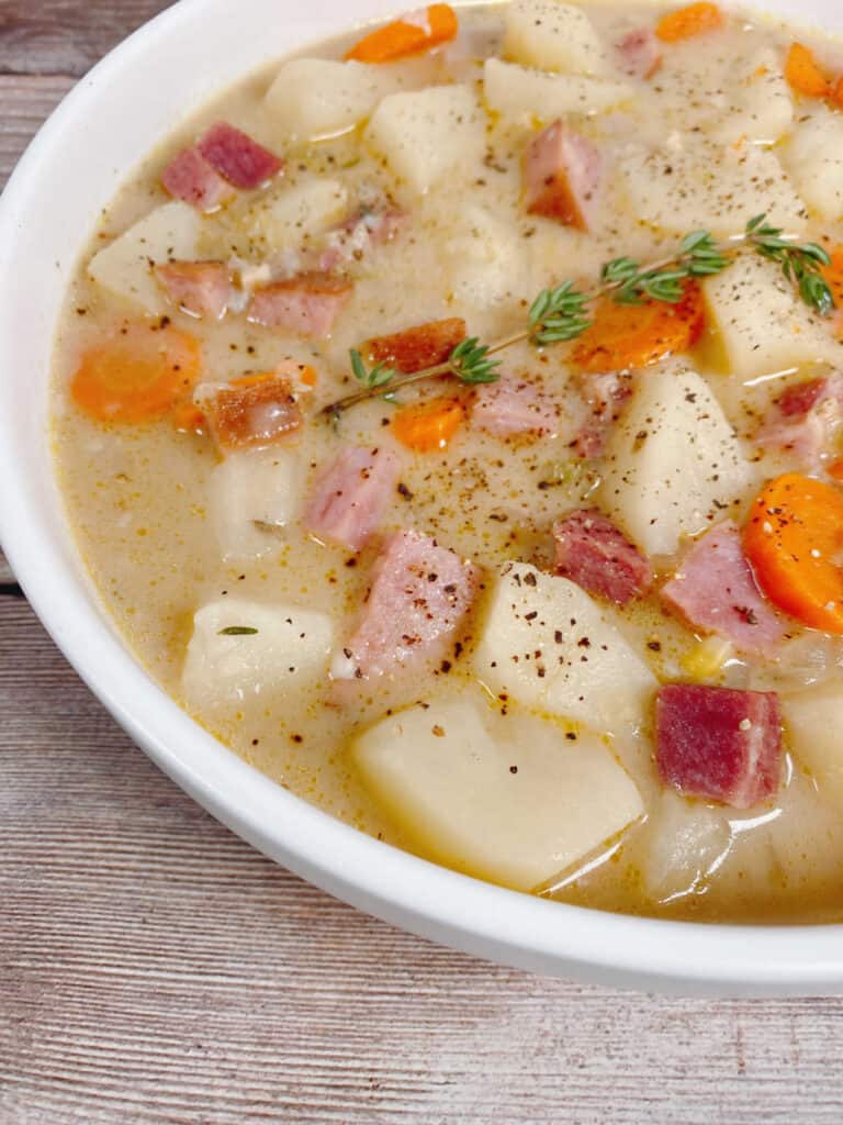 A large bowl of creamy ham and white soup takes over the entire image. A wooden background is barely visible around the bowl. Pieces of potato, ham, carrots and corn sit atop the creamy broth. The soup is seasoned with salt and pepper and a sprig of fresh thyme lays on top of the ingredients. 