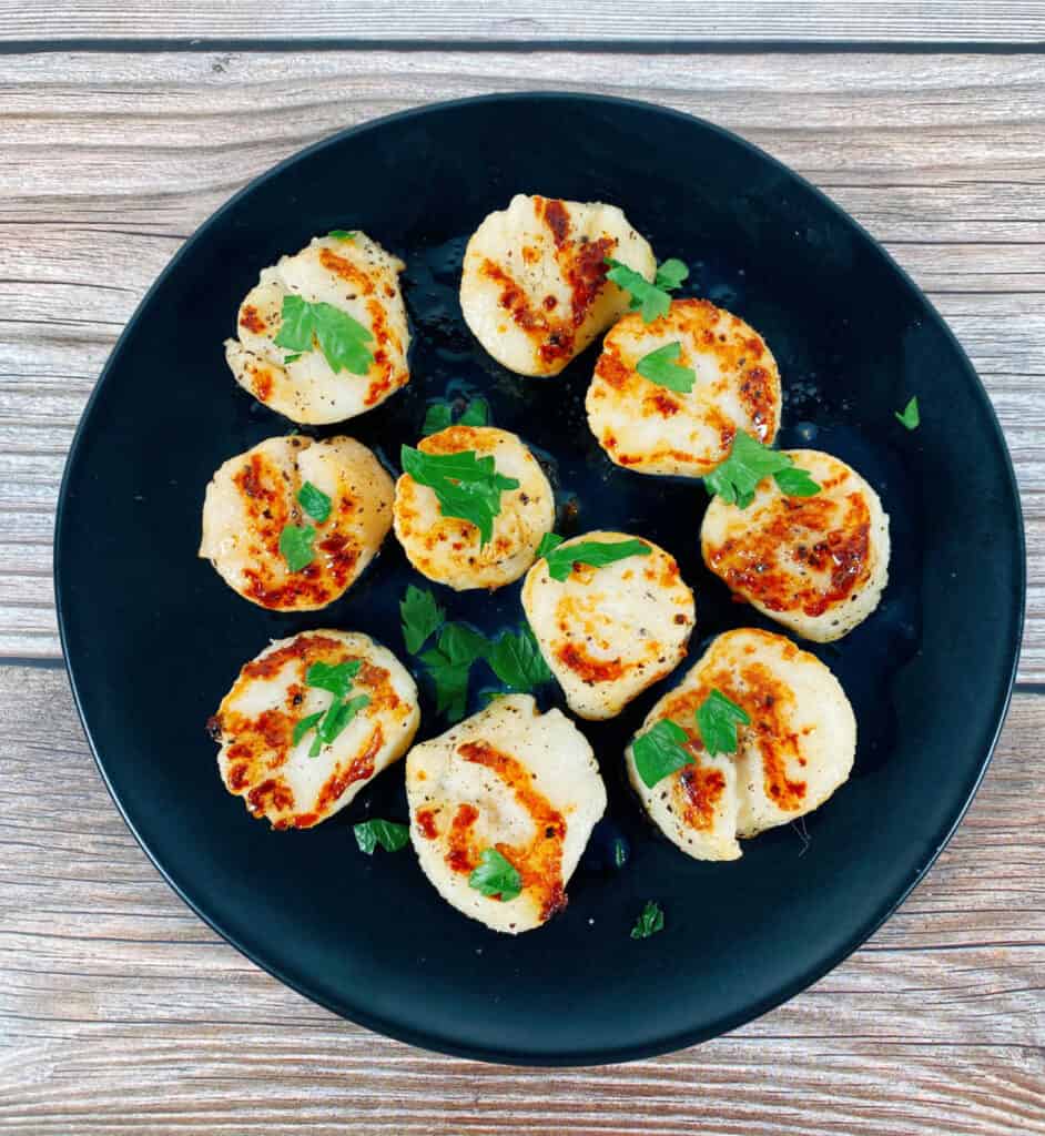 on a wooden background sits a round, black plate. Pan-seared scallops with golden tops sit on the plate and are sprinkled with fresh parsley. 