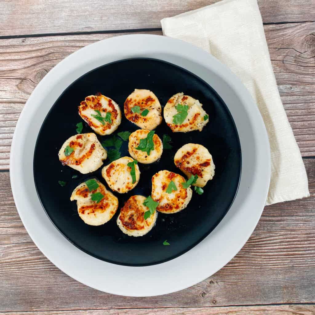 Pan Seared Scallops sprinkled with fresh parsley sit on a black plate stacked on top of a white plate.