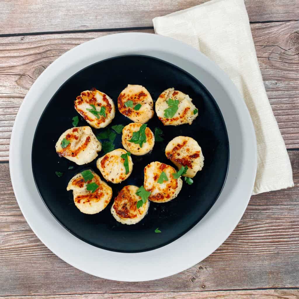 Pan Seared Scallops sprinkled with fresh parsley sit on a black plate stacked on top of a white plate. 