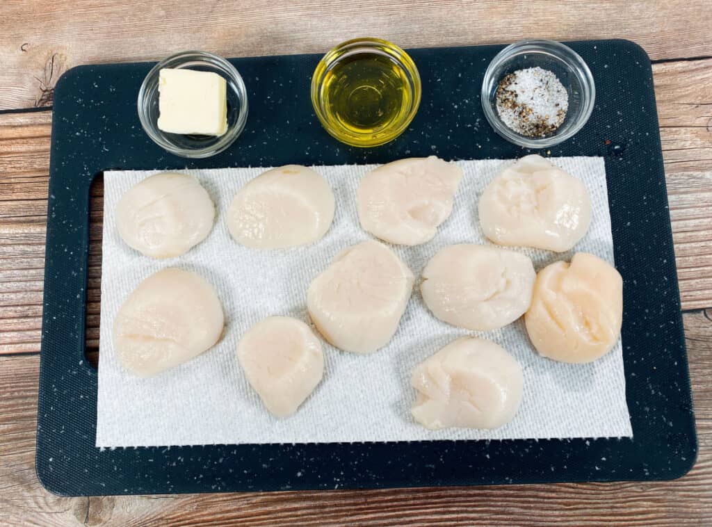 On a black cutting board sits the ingredients for pan seared scallops. Scallops are resting on a paper towel to remove excess water. Three dishes sit in front of the scallops. In the first is butter, in the second is olive oil and in the third is salt and pepper.