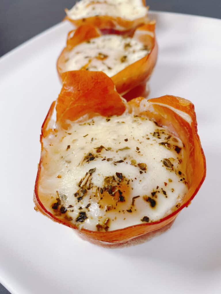 on a white plate sits two prosciutto egg cups - prosciutto is wrapped around the oven-baked egg topped with mozzarella cheese and Italian seasoning