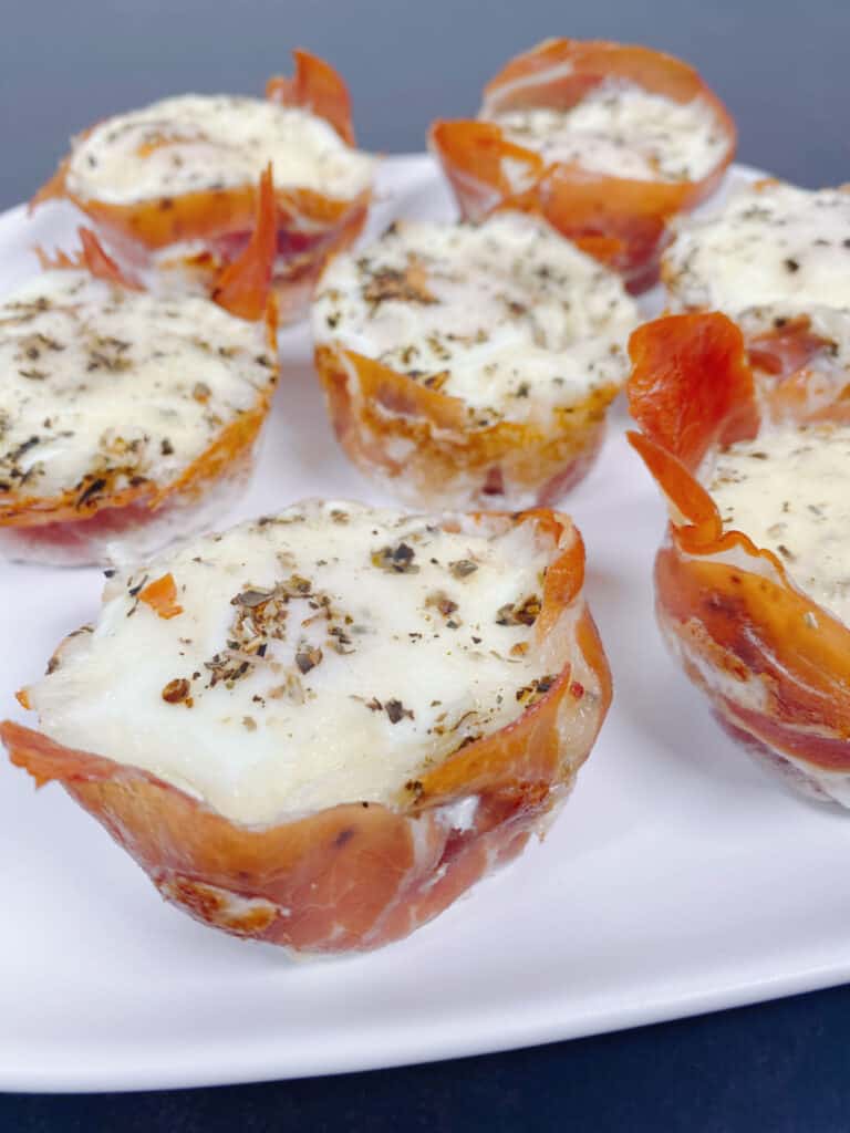 White plate of prosciutto egg cups - prosciutto wrapped around an oven-baked egg thats topped with mozzarella cheese and italian seasoning