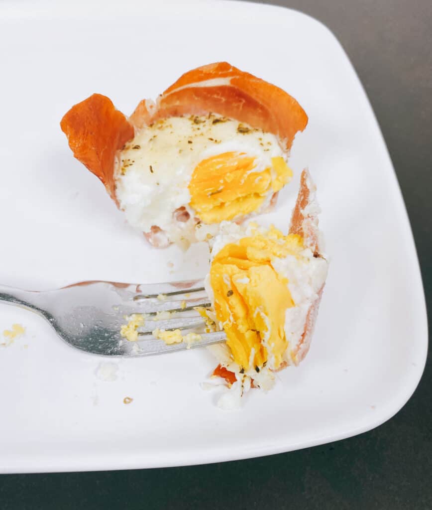 on a white plate sits a prosciutto egg cup cut open to show the soft boiled yolk inside. the top has melted mozzarella cheese and italian seasoning 