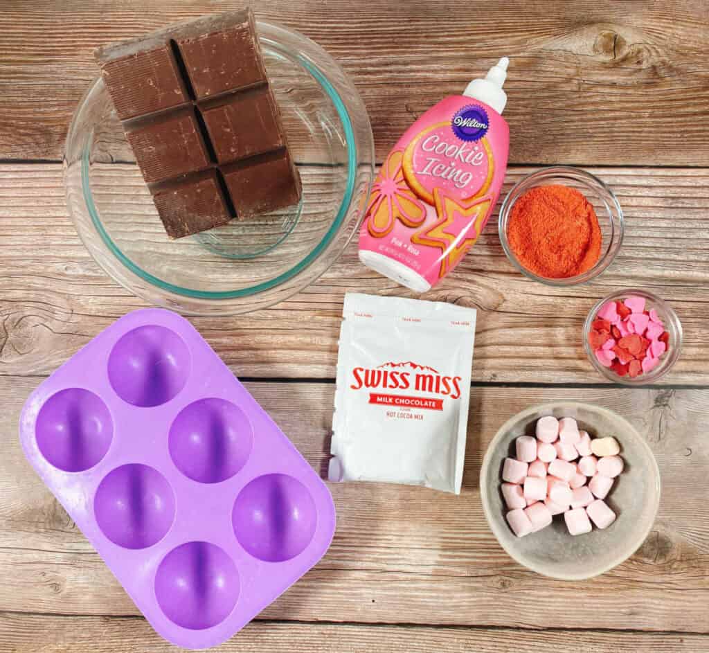 Ingredients for strawberry hot chocolate bombs sit on a wooden background. Ingredients include a silicone spherical mold, chocolate almond park, hot chocolate powder mix, powdered freeze dried strawberries, pink cookie icing. strawberry marshmallows and pink heart sprinkles. 