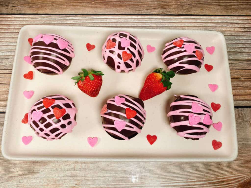 strawberry hot chocolate bombs sit on a pale pink rectangle platter on a wooden background. Bombs are decorated with pink cookie icing and heart sprinkles. On the plate are fresh strawberries and more heart sprinkles. 