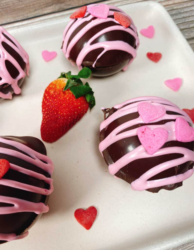 Spherical milk chocolate bombs, filled with hot cocoa powder, marshmallows and strawberry powder are decorated with pink icing and candy heart sprinkles. They sit on a pale pink rectangle platter surrounded by heart sprinkles and fresh strawberries. 