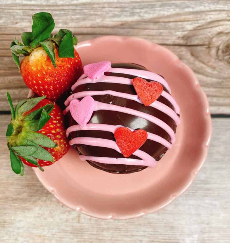 Close up image of a strawberry hot chocolate bomb sitting on a small pink stand with fresh strawberries. 