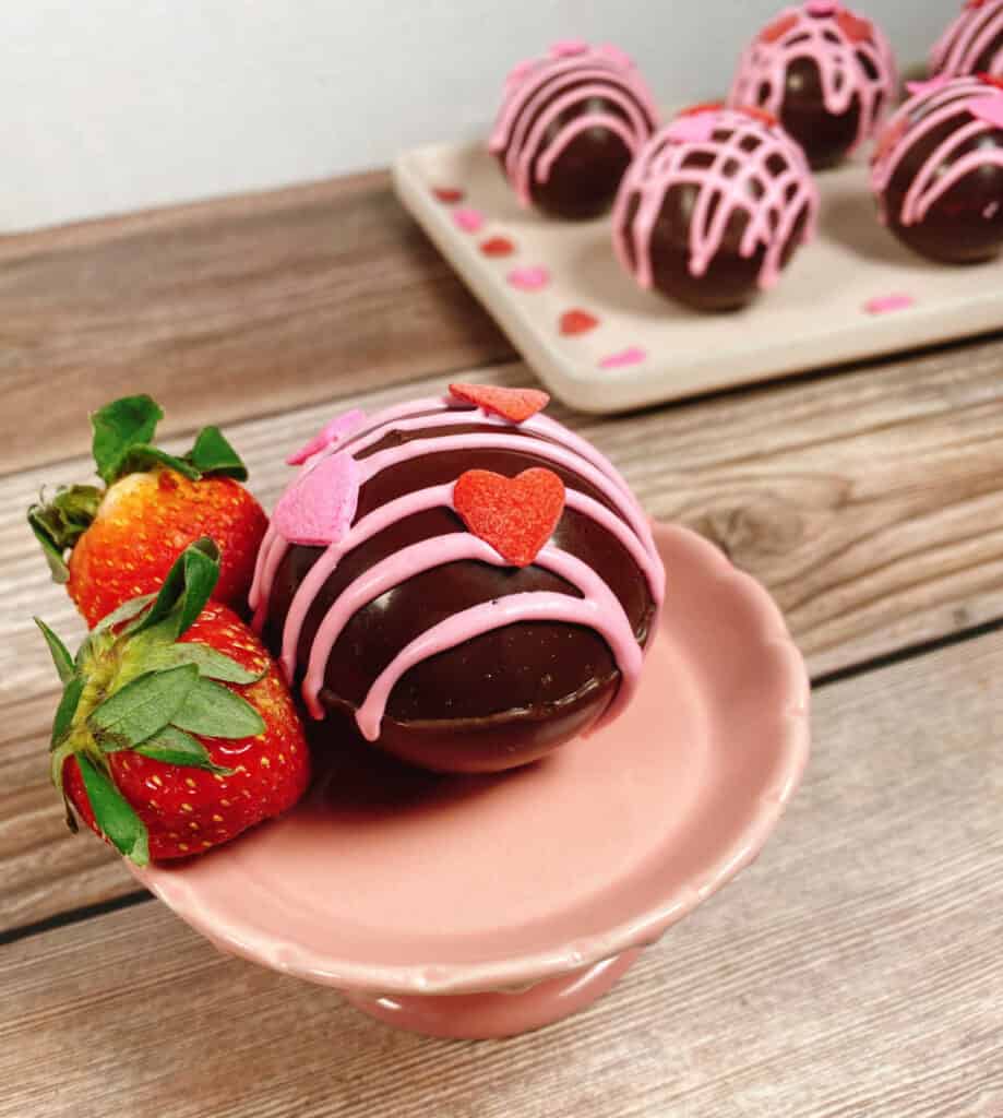 A strawberry hot chocolate bomb sits on a small pink stand with two strawberries. In the background, more hot chocolate bombs sit on a pale pink rectangle platter. 