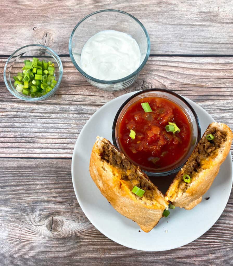 On a wooden background sits a glass bowls and a white plate. First bowl is full of chopped green onions. Second one is full of sour cream. On the white plate, there is a third bowl full o salsa. Also on the plate is an easy cheesy taco calzone, standing up against the bowl to show the meat and cheese in the inside.