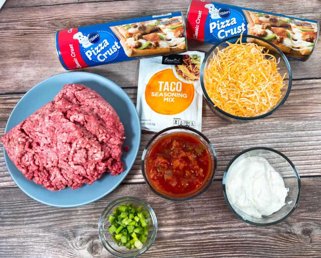 Ingredients for easy, cheesy taco calzones. Ingredients are ground beef, taco seasoning packet, shredded taco cheese, refrigerated pizza dough rolls, salsa, sour cream and green onions. 