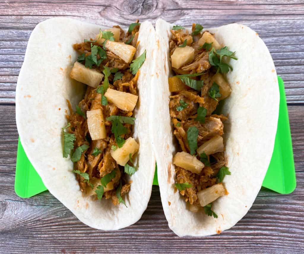 Two tacos sit in a taco stand on a wooden background. Flour tortillas are filled with the shredded, al pastor pork and are topped with pineapple, cilantro and onion. 