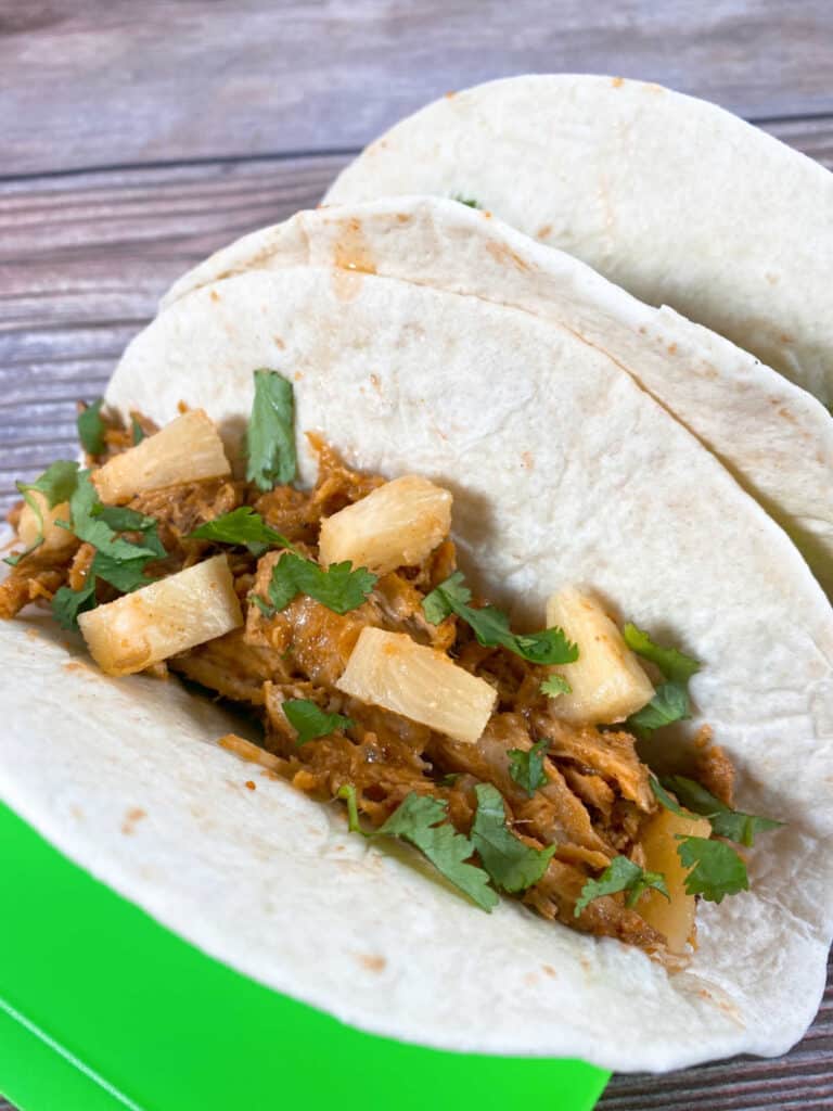 Two tacos sit in a taco stand on a wooden background. Flour tortillas are filled with the shredded, al pastor pork and are topped with pineapple, cilantro and onion. 