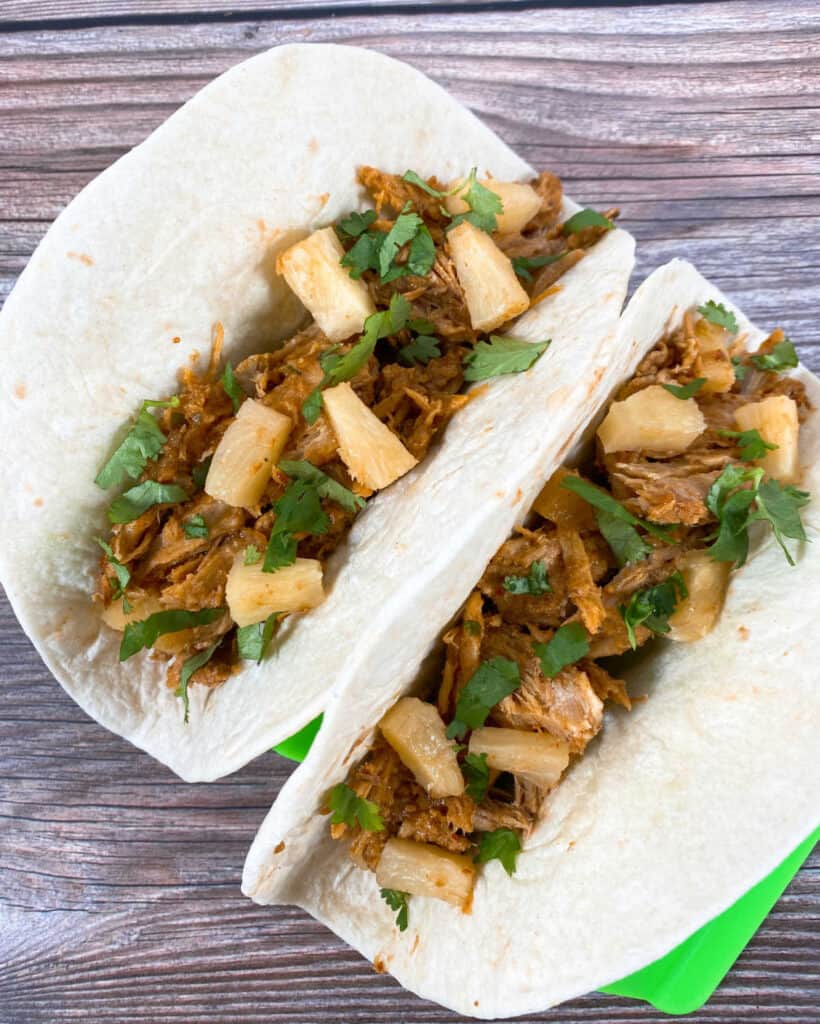 Two tacos sit in a taco stand on a wooden background. Flour tortillas are filled with the shredded, al pastor pork and are topped with pineapple, cilantro and onion. 