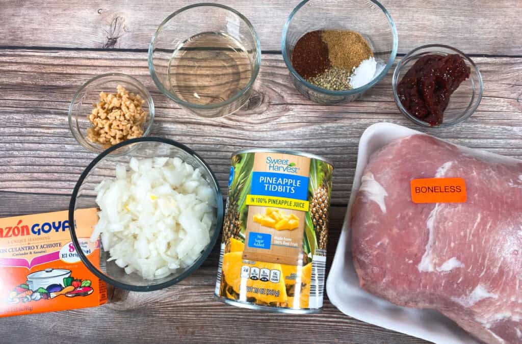 Ingredients for tacos al pastor sit in glass bowls on a wooden background. Ingredients are pork shoulder, canned pineapple, onion, vinegar, chiles in adobo, garlic, oregano, salt and Goya Sazon. To complete the meal, tortillas, cilantro and additional onion and pineapple are needed.