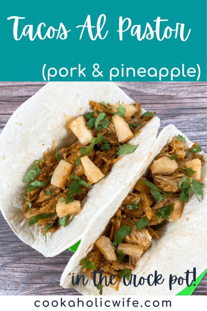On a wooden background sits two tacos at an angle. The flour tortillas are filled with Tacos Al Pastor, a marinated and shredded beef cooked in the slow cooker. The tacos are topped with pineapple, cilantro and onion.
