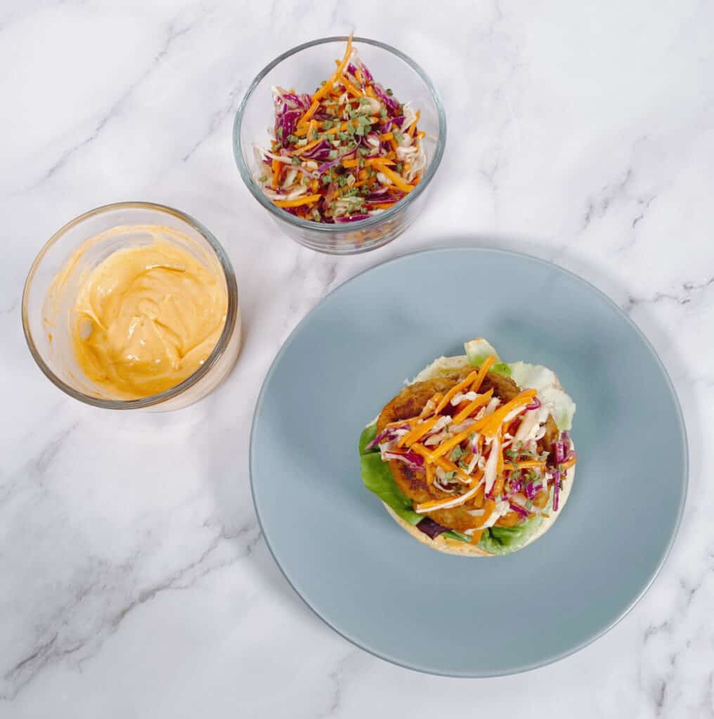 Marble background, blue plate. On the plate sits a thai turkey burger with the top part of the bun off. Colorful coleslaw tops the turkey burger with lettuce underneath and the bottom part of the bun. On the left sits two glass bowls, one with the spicy sriracha mayo and the other with the remaining Asian slaw. 