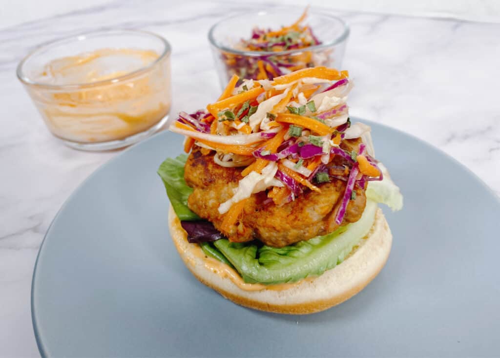 Marble background, blue plate. On the plate sits a thai turkey burger with the top part of the bun off. Colorful coleslaw tops the turkey burger with lettuce underneath and the bottom part of the bun. On the left sits two glass bowls, one with the spicy sriracha mayo and the other with the remaining Asian slaw. 