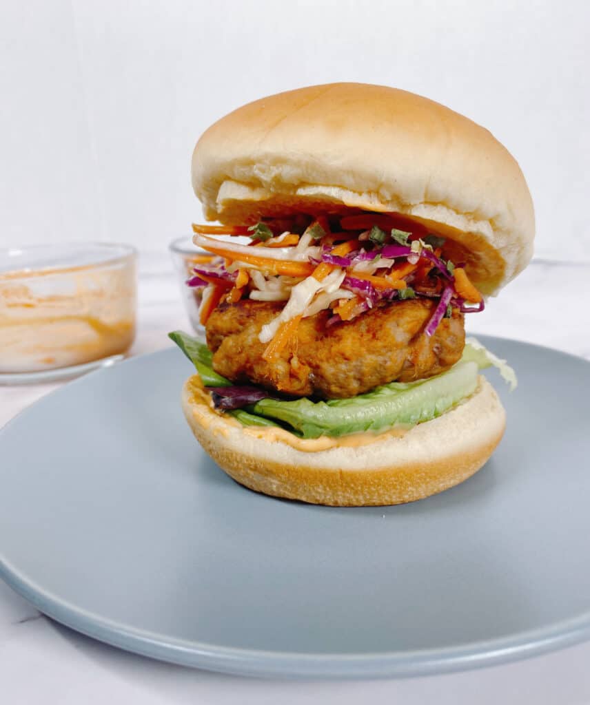 A Thai Turkey Burger stacked high with Asian slaw sits on a blue plate. In the background is a glass bowl of sriracha mayo that's slathered on both parts of the bun for extra flavor. 