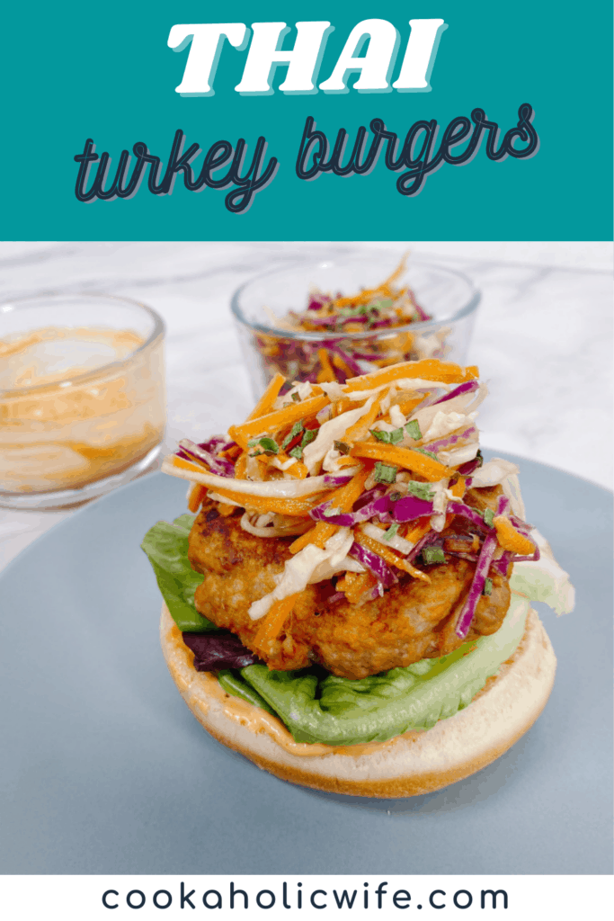 Thai Turkey Burgers: colorful asian slaw of carrots and cabbage sits on top of a thai-flavored turkey burger. Underneath is a bed of green leaf lettuce and the burger bun is slathered with a spicy sriracha mayo. All sitting on a blue plate. In the background are bowls with the asian slaw and spicy mayo. 