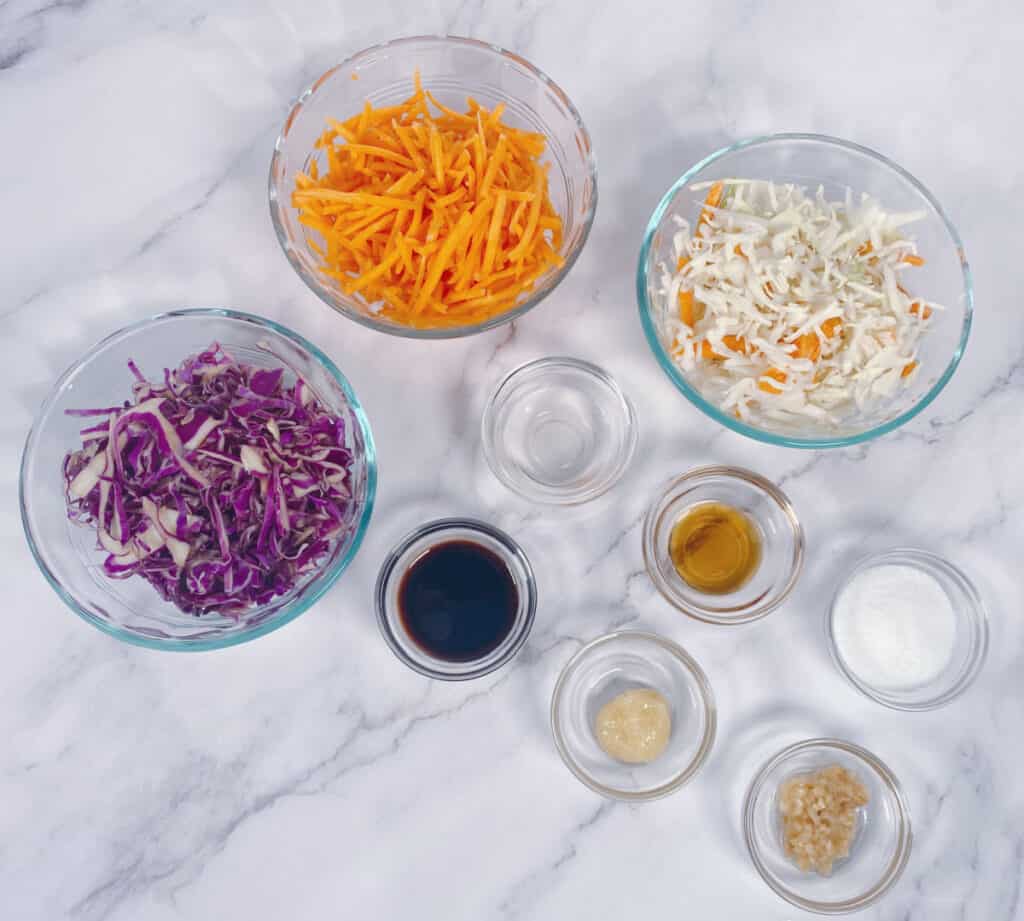 Ingredients for Asian Slaw to top the Thai Turkey Burgers sit in glass bowls on a marble background. Ingredients are green and purple cabbage, carrots, soy sauce, garlic, ginger, rice vinegar, sesame oil and sugar, 