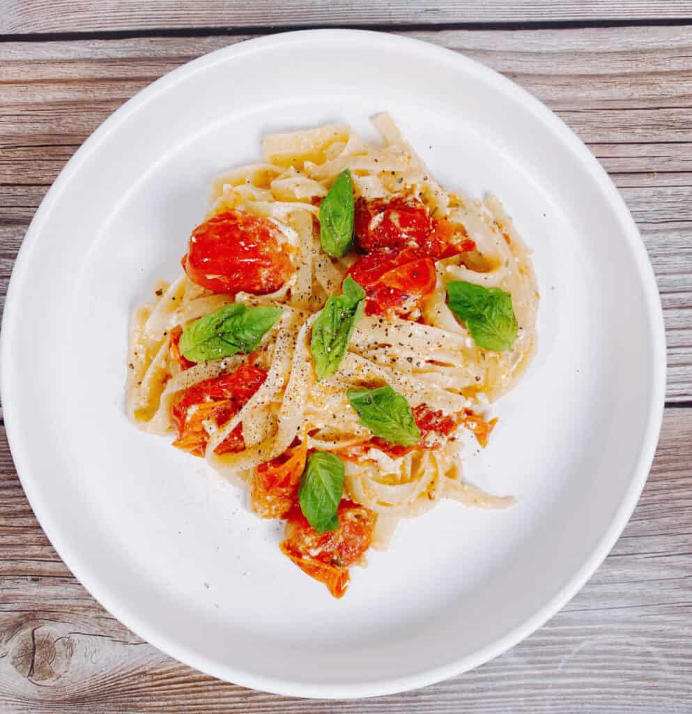 on a wooden background sits a white pasta plate. In the center of the plate is the baked feta tomato fettuccine, topped with fresh basil. 