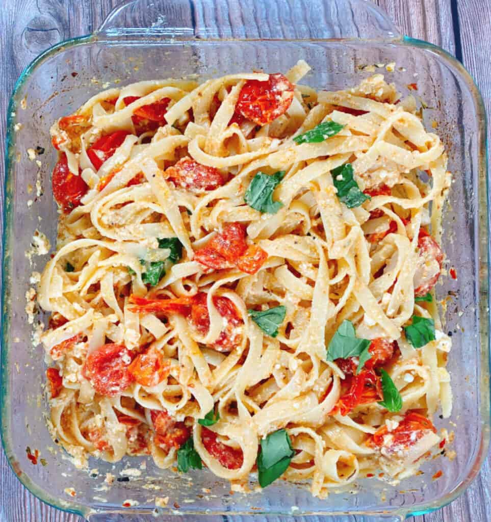 a glass baking dish full of baked feta tomato fettuccine sits on a wooden background. Fettuccine pasta is tossed with roasted tomatoes and feta cheese. Basil leaves top the dish.