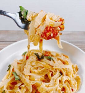 bowl full of baked feta tomato fettuccine on a wooden background. A fork holds up a bite of the pasta with tomato and basil.