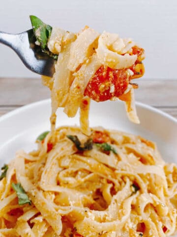 bowl full of baked feta tomato fettuccine on a wooden background. A fork holds up a bite of the pasta with tomato and basil.