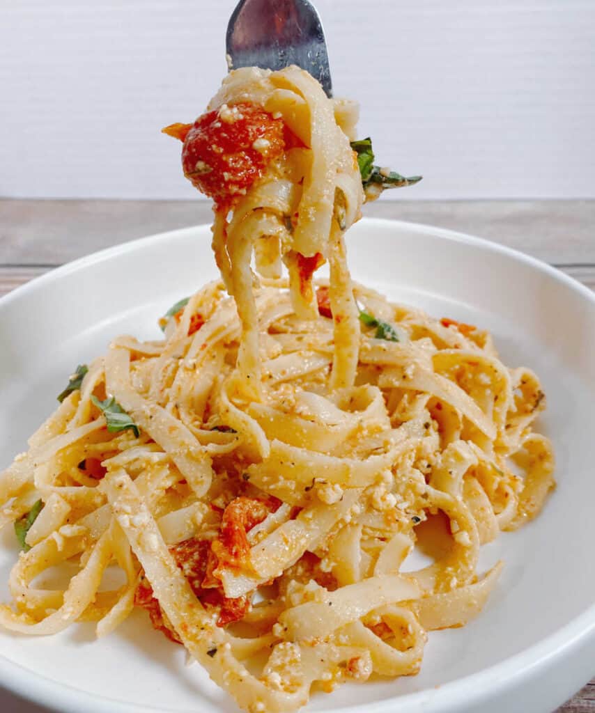 white pasta plate full of baked feta tomato fettuccine. Twisted on a fork held above the bowl is a roasted tomato and a basil leaf. 