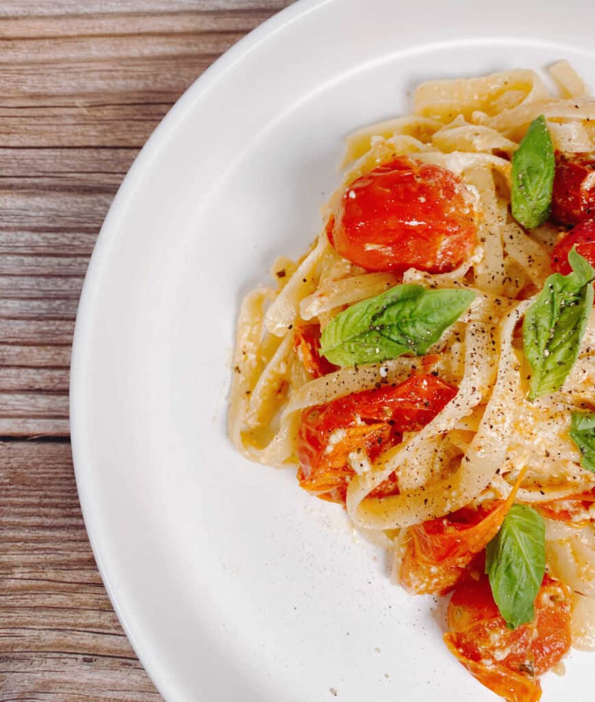 top view of dish, cut off on an angle. Wooden background, white plate. Baked feta tomato fettuccine takes over the right side of the image, topped with fresh basil leaves. 