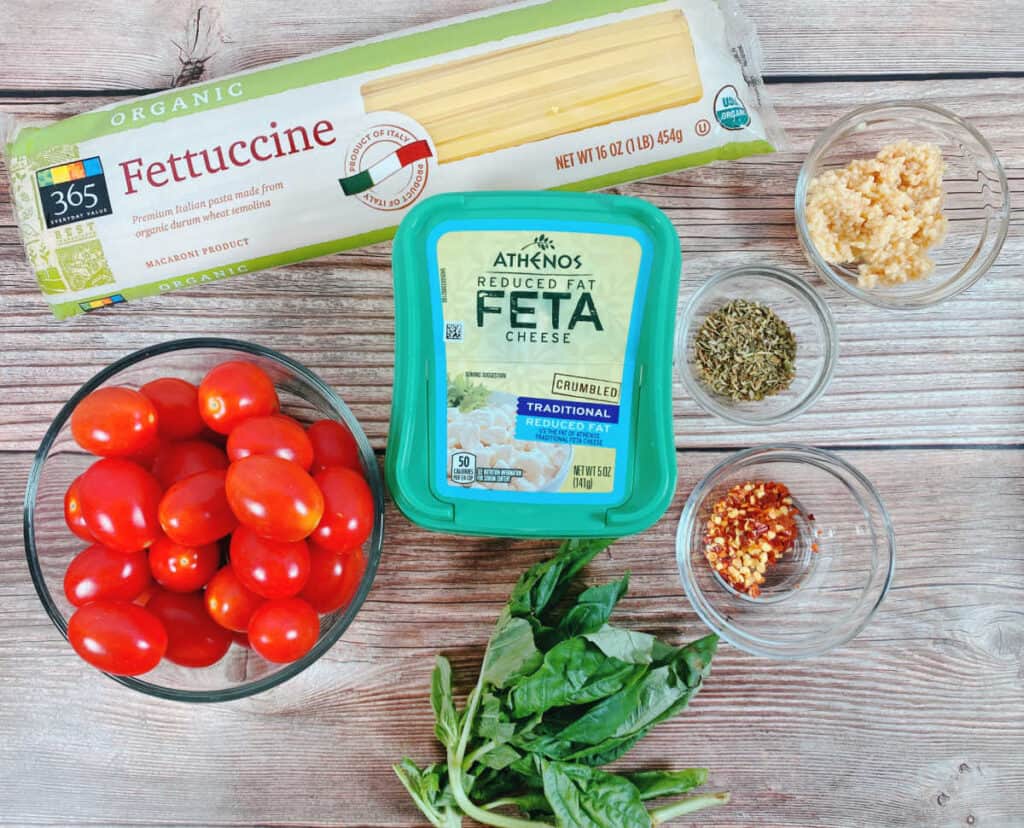 ingredients for baked feta tomato fettuccine sit on a wooden background. Fettuccini pasta, grape tomatoes, feta cheese, italian seasoning, garlic, basil, crushed red pepper. Not pictured: olive oil.