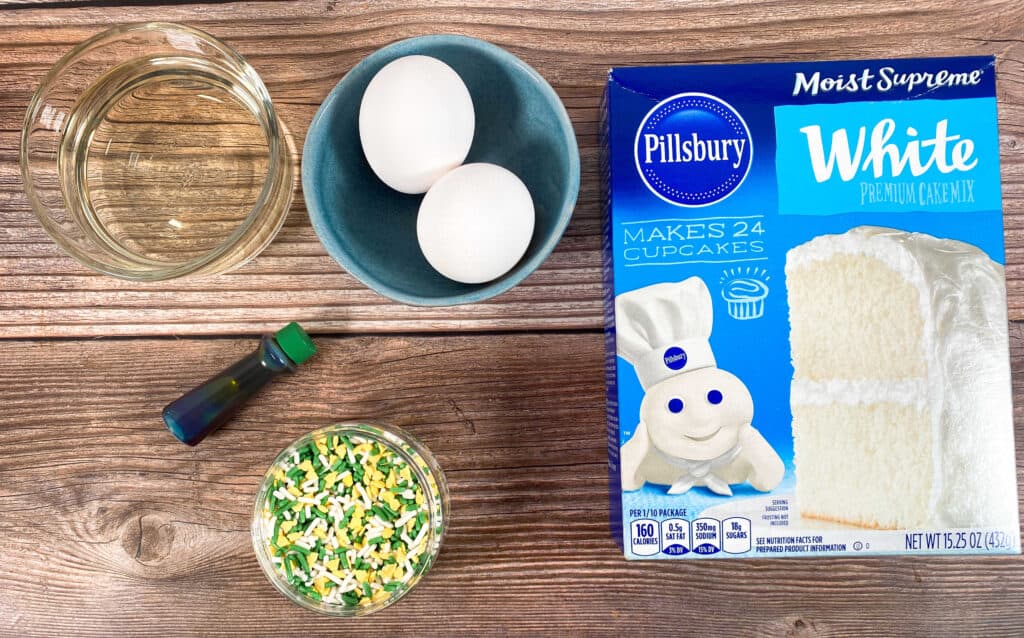 ingredients for irish cake mix cookies sit on a wooden background - cake mix, vegetable oil, eggs, green food coloring and sprinkles. 