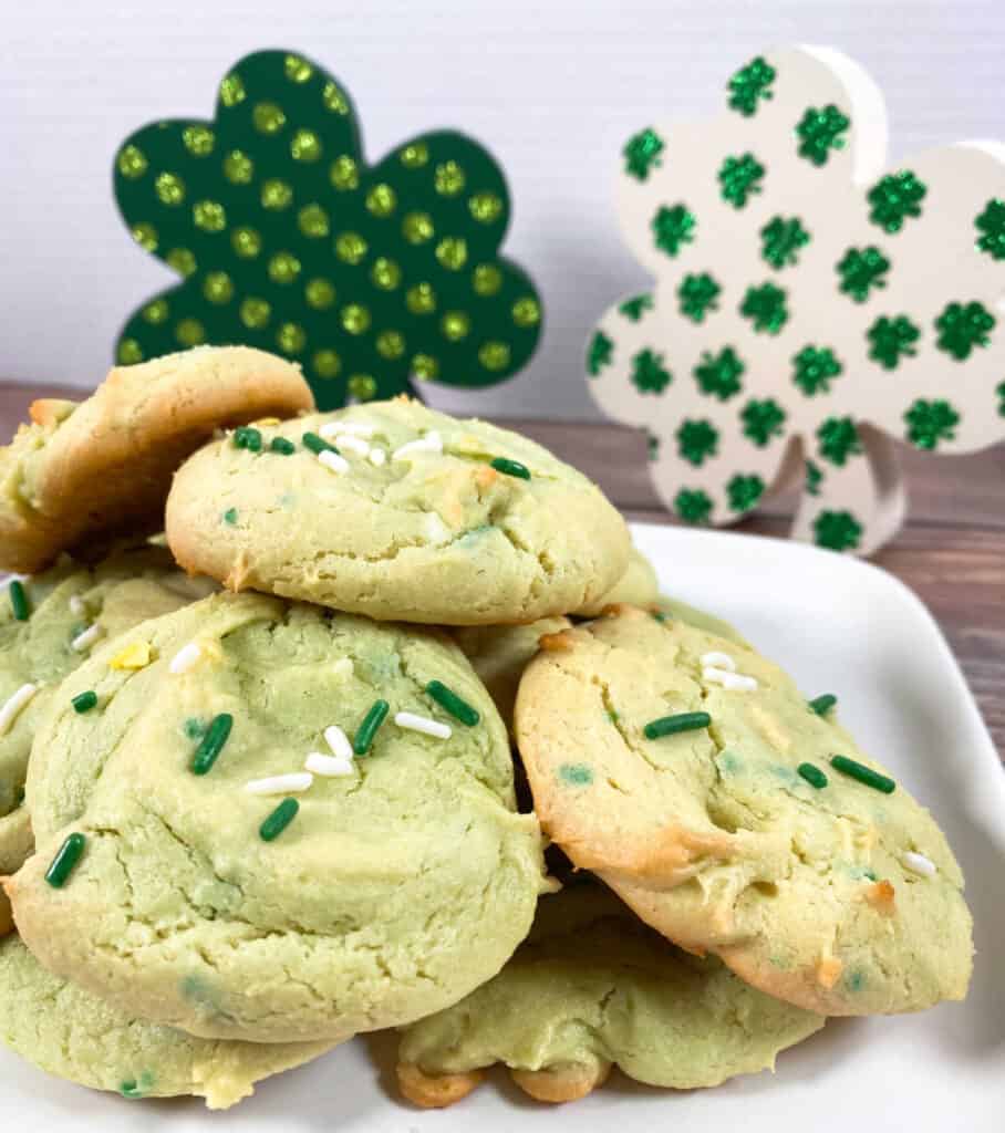 green hued cake mix cookies topped with sprinkles sit on a white plate with white and green wooden shamrock decorations behind them. 