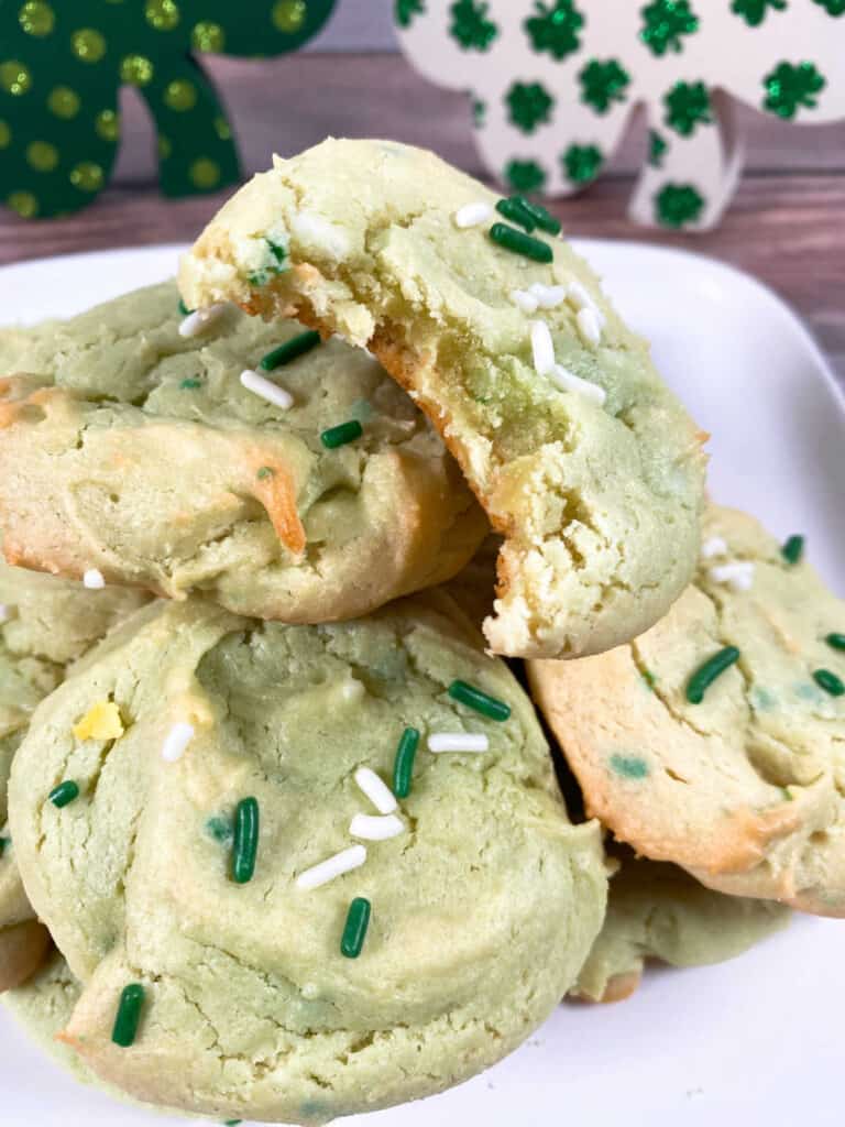 on a white plate sits irish cake mix cookies. cookies are tinted green and have green and white sprinkles on them. cookies are piled up high and the top cookie has a bite missing. 