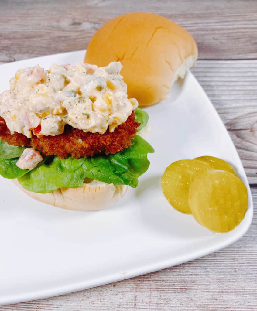 Open-faced view of the sandwich, with a heaping pile of the creamy corn relish sitting on top a piece of crispy, fried pork on a burger bun. Pickles sit off to the side of the sandwich. 