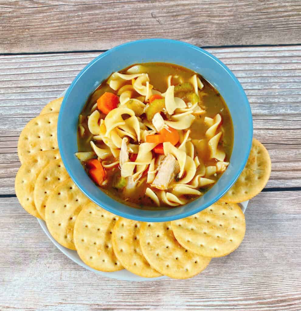 A hearty bowl of chicken noodle soup, full of shredded chicken, carrots and celery sits surrounded by crackers on a wooden background. 