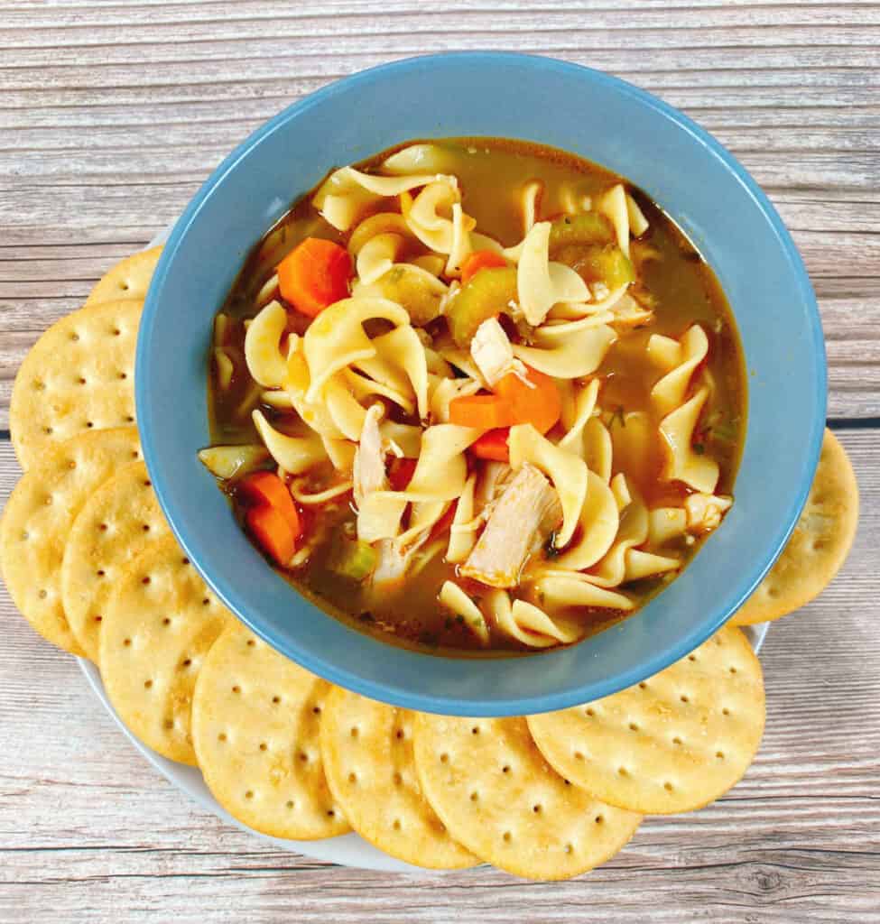 close up bowl of chicken noodle soup, with egg noodles, shredded chicken, carrots and celery visible. Round crackers surround the bowl and it's sitting on a wooden background. 