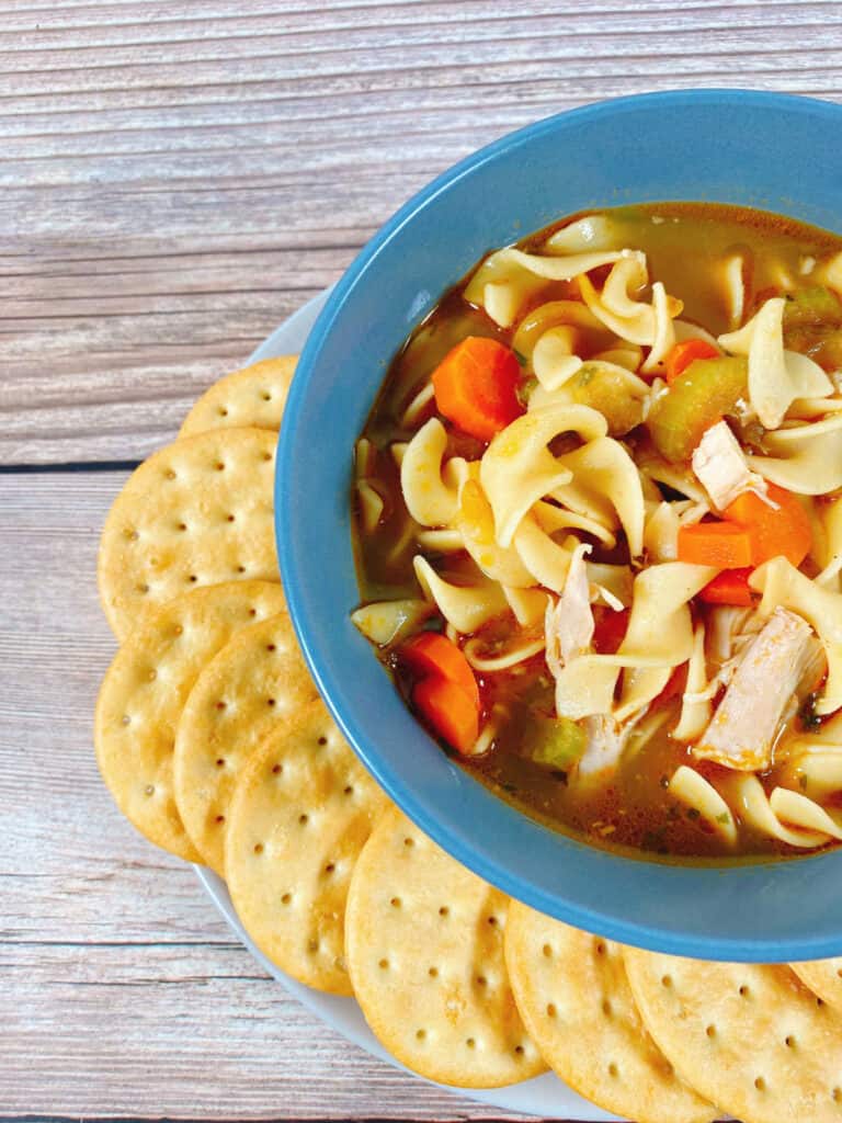 Side image of chicken noodle soup - close up of the soup, showing the carrots, noodles and chicken. Crackers sit next to the bowl. 