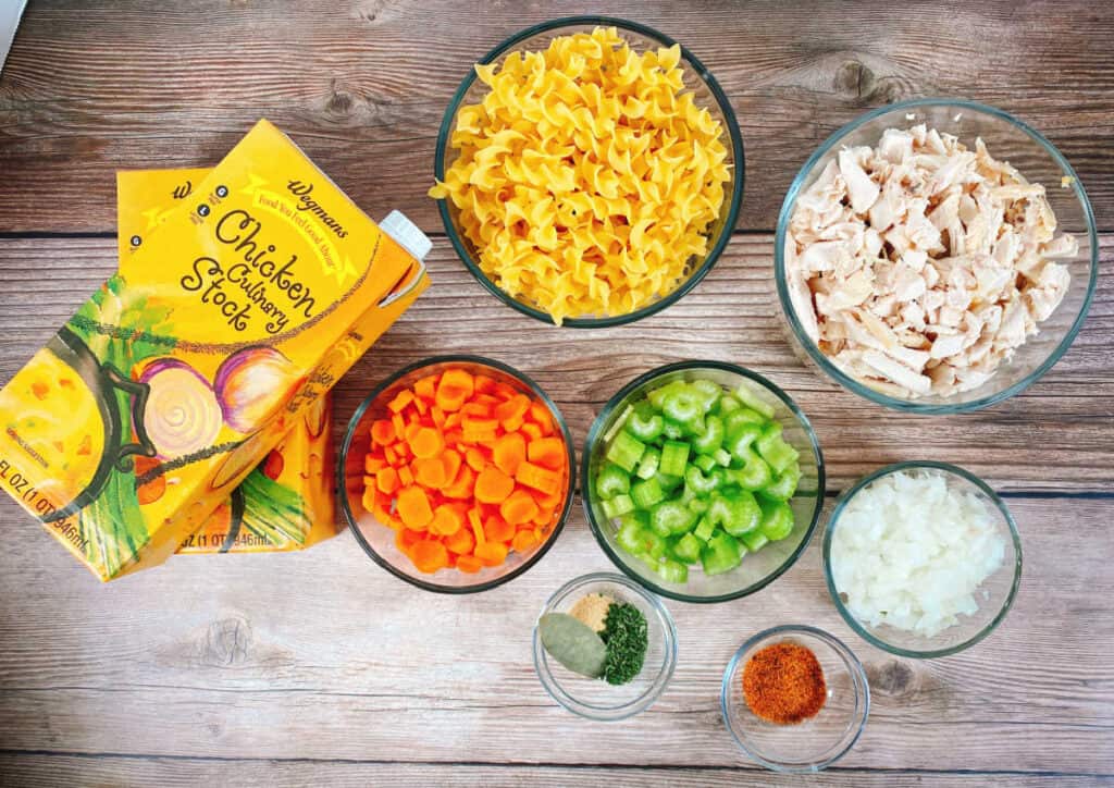 Ingredients for Quick and Easy Chicken Noodle Soup sit on a wooden background. From left to right - two cartons of chicken stock, egg noodles, carrots, celery, seasonings, onion and shredded chicken. 