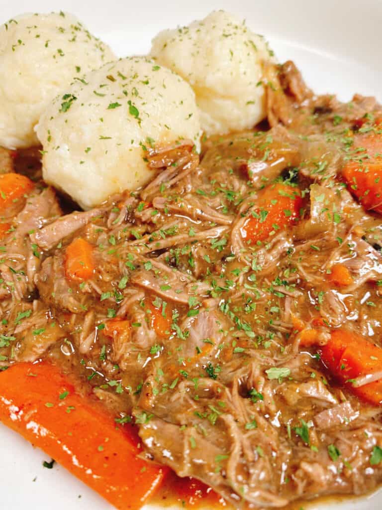 Close up picture of the sour beef and dumplings, the beef is shredded and mix in with the carrots and onions. The potato dumplings are in the back and the entire dish is sprinkled with fresh parsley.