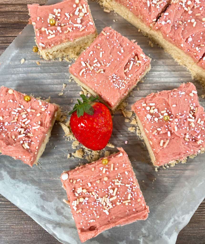 strawberries and cream cookie bars are cut into squares sitting on a piece of parchment paper on a wooden background. Frosting on the bars is pink and covered in sprinkles. a strawberry sits in the center of the bars. 