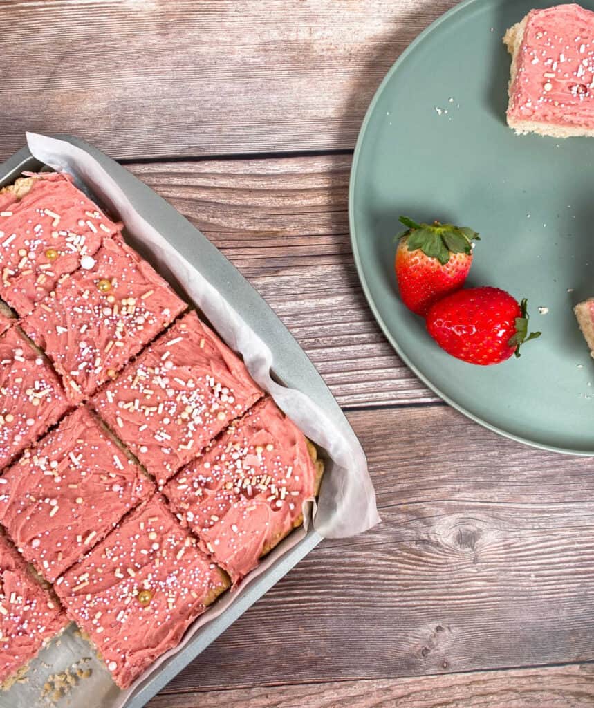 on the left side of the image is strawberries and cream cookie bars in a baking dish, cut into squares. On the right are strawberries and some of the cut up cookie bar on a mint green plate. 