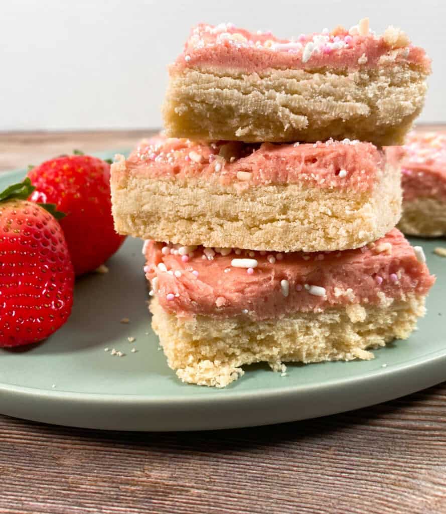 strawberries and cream cookie bars are stacked 3 high on a mint green plate on a wooden background. on the left are two fresh strawberries. 