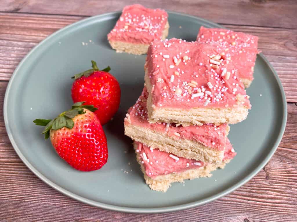 on a wooden background sits a mint green plate. three strawberries and cream cookie bars are stacked on top of each other and two more are in the background. two strawberries sit on the left side of the plate. 