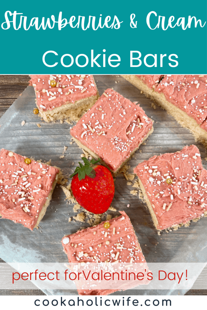 strawberries and cream cookie bars sit on a sheet of parchment paper with a strawberry in the center. bars are decorated with pink, strawberry frosting and sprinkles. 