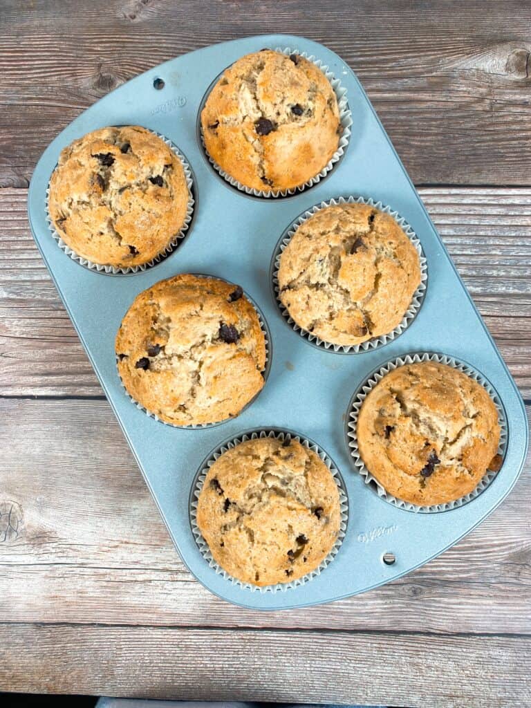 image looking down on a tray of baked jumbo banana muffins with tall domes rising way over the muffin pan. Muffin pan sits on a wooden background. 