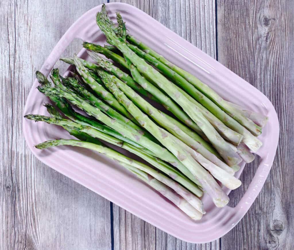 spears of asparagus sit on a pale pink platter on a wooden background