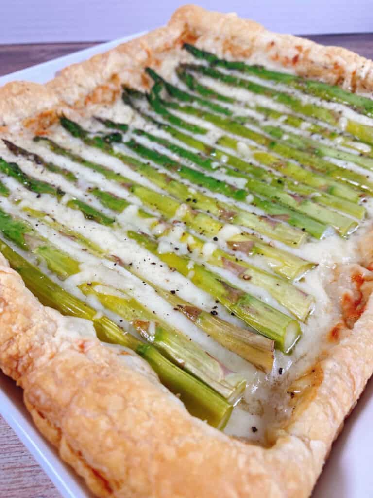 Close up image of asparagus and gruyere tart. Puff pastry is layered with a sauce and shredded gruyere cheese and topped with fresh asparagus spears, then baked to golden perfection. 